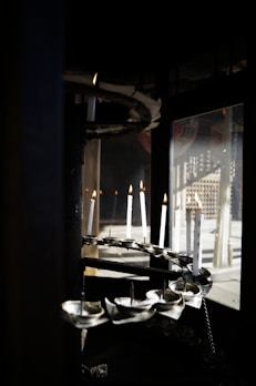 Several white candles are positioned in a spiral formation within a dark, enclosed space. They are lit, casting a warm glow, and there are reflections visible in a glass window nearby.