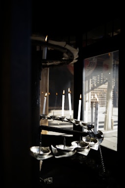 Several white candles are positioned in a spiral formation within a dark, enclosed space. They are lit, casting a warm glow, and there are reflections visible in a glass window nearby.