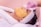 A person is lying back with a yellow facial mask being applied using a spatula. They are wearing a white headband and a white robe. The person applying the mask is wearing pink gloves.