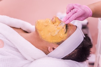 A person is lying back with a yellow facial mask being applied using a spatula. They are wearing a white headband and a white robe. The person applying the mask is wearing pink gloves.