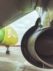 A close-up view of an aircraft's engine and wing with another airplane in the background. The aircraft in the background displays the S7 Airlines branding in green colors, parked on the concrete surface of an airport runway under an overcast sky.