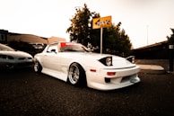 A modified white sports car with pop-up headlights is parked on an asphalt surface. The car has a red banner across the top of the windshield and is fitted with wide wheels and flared fenders. Nearby, another car is partially visible. The background features a yellow sign with graffiti, trees, and a concrete structure under a clear sky.