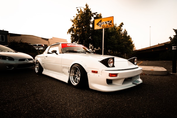 A modified white sports car with pop-up headlights is parked on an asphalt surface. The car has a red banner across the top of the windshield and is fitted with wide wheels and flared fenders. Nearby, another car is partially visible. The background features a yellow sign with graffiti, trees, and a concrete structure under a clear sky.