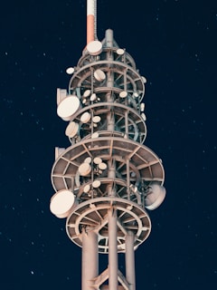 A tall communication tower with multiple satellite dishes and antennas arranged in tiers against a clear night sky. The structure is illuminated with artificial lighting, creating a contrast with the dark background, and stars can be faintly seen.