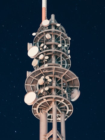 A tall communication tower with multiple satellite dishes and antennas arranged in tiers against a clear night sky. The structure is illuminated with artificial lighting, creating a contrast with the dark background, and stars can be faintly seen.
