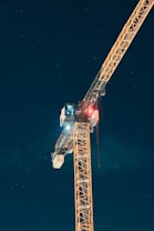 A tall construction crane is illuminated against a night sky filled with stars. The crane is yellow and features a cabin with a few lights, and there are visible structural elements that are intricately detailed. The sky is dark, creating a striking contrast with the brightly lit crane.