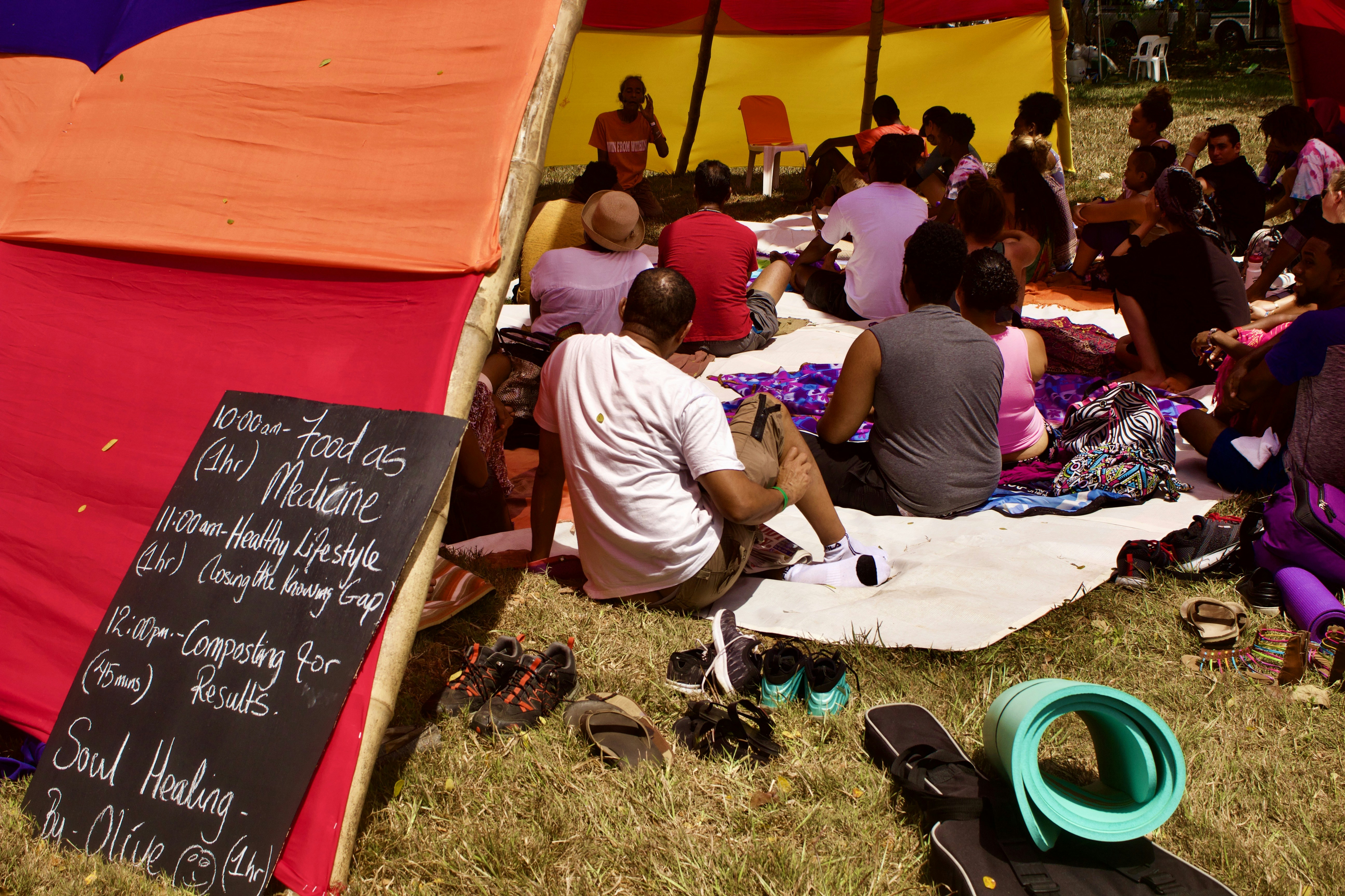A group of people sits under a colorful tent made of red, yellow, and orange fabric. They appear to be attending a workshop or class. A chalkboard on the left lists a schedule with topics like 'Food as Medicine' and 'Healthy Lifestyle.' The participants sit on mats and blankets, listening to someone speaking at the front. The atmosphere is casual and relaxed with shoes and personal items placed on the grass nearby.