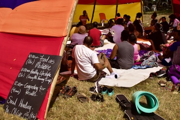 A group of people sits under a colorful tent made of red, yellow, and orange fabric. They appear to be attending a workshop or class. A chalkboard on the left lists a schedule with topics like 'Food as Medicine' and 'Healthy Lifestyle.' The participants sit on mats and blankets, listening to someone speaking at the front. The atmosphere is casual and relaxed with shoes and personal items placed on the grass nearby.