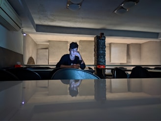 A person sits alone in a dimly lit room with a minimalist design, looking at a phone with a contemplative expression. The room features geometric wall patterns and a reflective surface in the foreground. Some furniture and an air conditioning unit are visible.