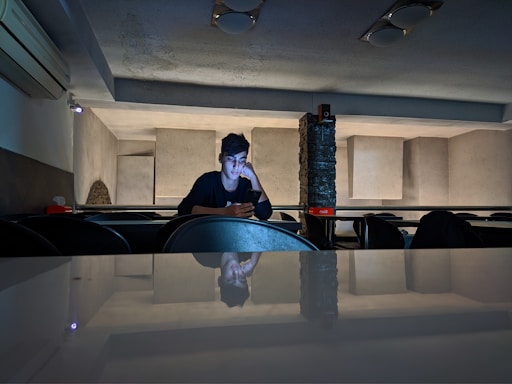 A person sits alone in a dimly lit room with a minimalist design, looking at a phone with a contemplative expression. The room features geometric wall patterns and a reflective surface in the foreground. Some furniture and an air conditioning unit are visible.