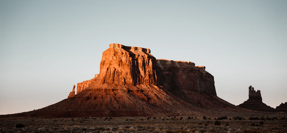 The image depicts a desert landscape with a large, light brown rock formation dominating the center of the frame. The rock formation is surrounded by a vast expanse of dry, barren land that stretches out to meet a clear blue sky at the horizon. In the distance, several smaller rocks can be seen scattered across the scene.