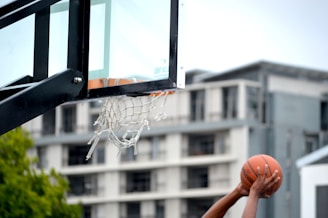A basketball is being held by a person, aimed towards a hoop with a net in an outdoor setting. The background shows a building with multiple floors, and there are some trees visible.