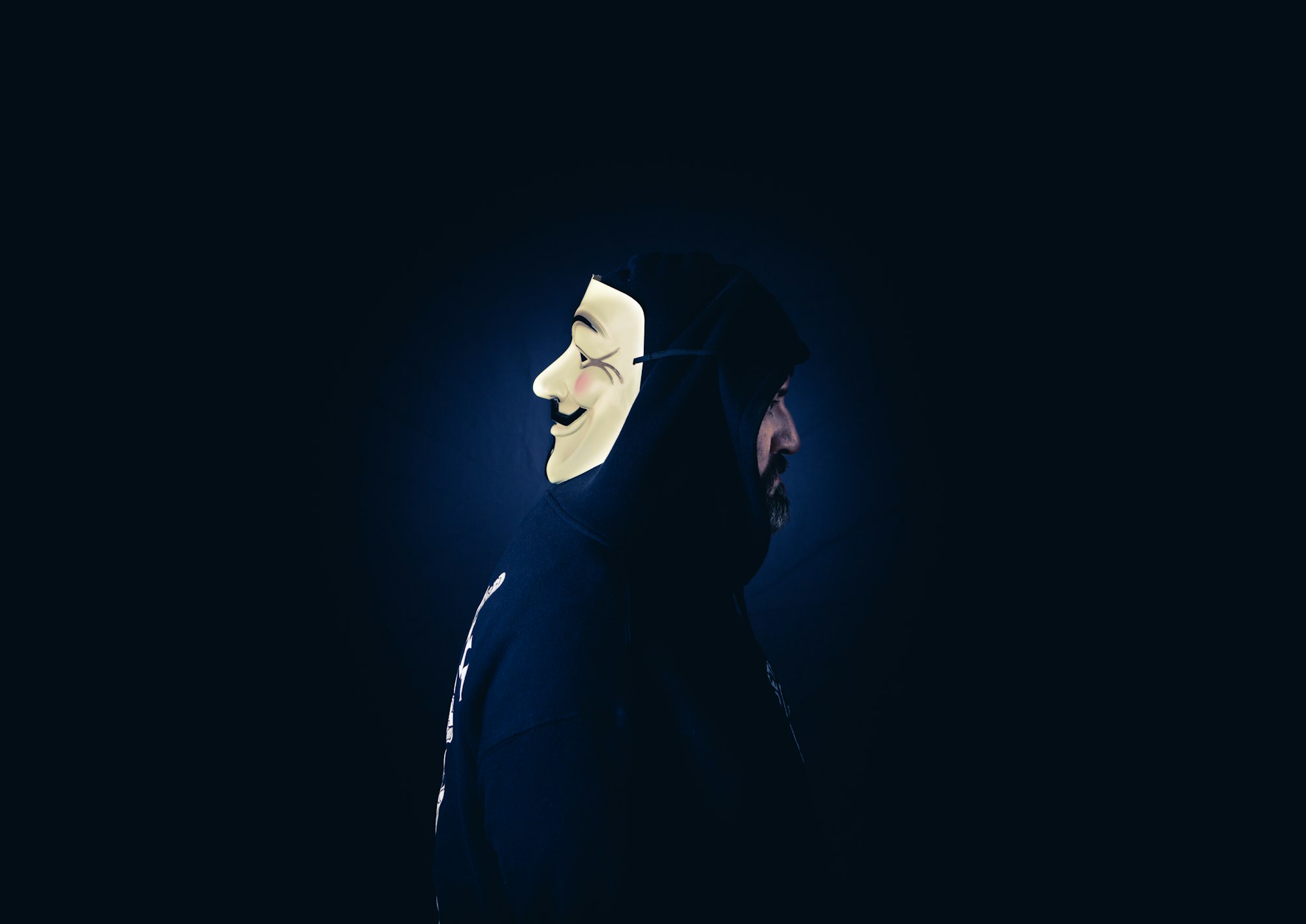 A man in a hoodie wears a Guy Fawkes mask on the back of head. 