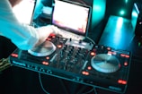 A person is operating a DJ controller with two turntables and various control knobs. A laptop is open in the background, possibly providing a digital music interface. The scene is illuminated with bright LED lights, giving it a vibrant and dynamic atmosphere.