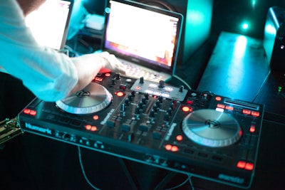 A person is operating a DJ controller with two turntables and various control knobs. A laptop is open in the background, possibly providing a digital music interface. The scene is illuminated with bright LED lights, giving it a vibrant and dynamic atmosphere.