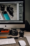 A workspace setup featuring a desktop computer with a screen displaying photo editing software. In front of the monitor, various camera equipment including a digital camera, camera lens, and memory cards are placed on a desk. A keyboard and mouse are also visible, along with some lens caps and an external hard drive.