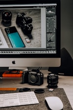 A workspace setup featuring a desktop computer with a screen displaying photo editing software. In front of the monitor, various camera equipment including a digital camera, camera lens, and memory cards are placed on a desk. A keyboard and mouse are also visible, along with some lens caps and an external hard drive.
