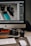 A workspace setup featuring a desktop computer with a screen displaying photo editing software. In front of the monitor, various camera equipment including a digital camera, camera lens, and memory cards are placed on a desk. A keyboard and mouse are also visible, along with some lens caps and an external hard drive.