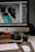 A workspace setup featuring a desktop computer with a screen displaying photo editing software. In front of the monitor, various camera equipment including a digital camera, camera lens, and memory cards are placed on a desk. A keyboard and mouse are also visible, along with some lens caps and an external hard drive.