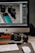 A workspace setup featuring a desktop computer with a screen displaying photo editing software. In front of the monitor, various camera equipment including a digital camera, camera lens, and memory cards are placed on a desk. A keyboard and mouse are also visible, along with some lens caps and an external hard drive.