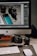 A workspace setup featuring a desktop computer with a screen displaying photo editing software. In front of the monitor, various camera equipment including a digital camera, camera lens, and memory cards are placed on a desk. A keyboard and mouse are also visible, along with some lens caps and an external hard drive.