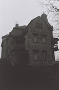 A large, old, and possibly abandoned wooden house with a dark and eerie appearance. The building shows signs of wear, with boarded windows and a bleak atmosphere. Bare tree branches are visible, adding to the ominous setting.