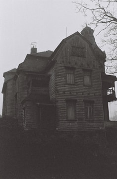 A large, old, and possibly abandoned wooden house with a dark and eerie appearance. The building shows signs of wear, with boarded windows and a bleak atmosphere. Bare tree branches are visible, adding to the ominous setting.
