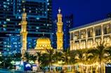 A well-lit mosque with multiple minarets, intricate architectural details, and a large central dome. The mosque is surrounded by palm trees and buildings, with a backdrop of modern high-rise structures. The night setting enhances the warm golden lights illuminating the mosque and nearby building.