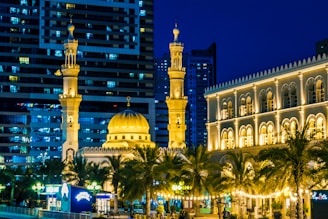 A well-lit mosque with multiple minarets, intricate architectural details, and a large central dome. The mosque is surrounded by palm trees and buildings, with a backdrop of modern high-rise structures. The night setting enhances the warm golden lights illuminating the mosque and nearby building.