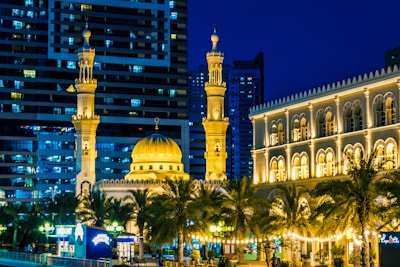 A well-lit mosque with multiple minarets, intricate architectural details, and a large central dome. The mosque is surrounded by palm trees and buildings, with a backdrop of modern high-rise structures. The night setting enhances the warm golden lights illuminating the mosque and nearby building.