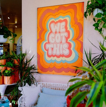 A colorful poster with the phrase 'We Got This' sits amidst vibrant green plants. The poster features bold, wavy patterns in orange, yellow, and blue tones, contributing to a warm and optimistic atmosphere. The surrounding environment includes potted plants and a light-colored wall, adding to the lively, indoor garden feel.