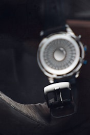 A close-up view of a stylish wristwatch with a unique circular design, featuring intricate patterns on the watch face and a black leather strap with a silver buckle. The lighting accentuates the metallic elements, while the background is dark and slightly blurred.