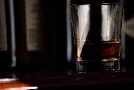 A glass filled with a dark amber liquid is placed on a polished surface, with dimly lit bottles forming a blurred background. The lighting highlights the contours and reflections on the glass, creating a sophisticated and moody atmosphere.