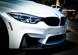 A close-up of the front end of a white BMW car, highlighting its sleek contours and distinctive grille. The headlights with their intricate design are prominently visible, as well as the car's sporty bumper.