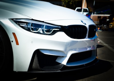 A close-up of the front end of a white BMW car, highlighting its sleek contours and distinctive grille. The headlights with their intricate design are prominently visible, as well as the car's sporty bumper.