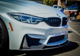 A close-up view of the front of a white BMW car featuring its distinctive grille, sleek design, and modern headlights. The setting appears to be an outdoor automotive event with other vehicles and equipment visible in the blurred background.