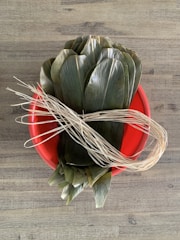 A red plastic basin contains a bundle of large green leaves and pieces of dried straw or twine, placed on a wooden surface.
