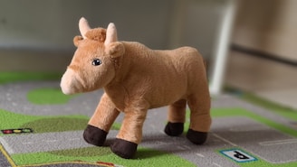 A plush toy resembling a brown bull with light brown fur and dark brown hooves stands on a green and gray textured play mat. The toy has small horns and a cute facial expression.