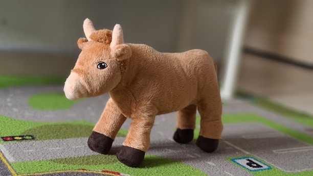 A plush toy resembling a brown bull with light brown fur and dark brown hooves stands on a green and gray textured play mat. The toy has small horns and a cute facial expression.