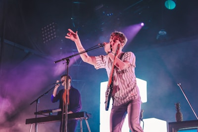 A musical performance on stage featuring two musicians. The foreground musician is energetically singing into a microphone, while playing a guitar, dressed in a striped shirt and jeans. In the background, another musician plays a keyboard. The stage is lit with vibrant purple and blue lights, creating a dynamic atmosphere. A pineapple is oddly placed on a speaker nearby.