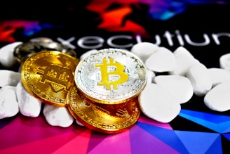 Several physical Bitcoin coins, displaying their iconic 'B' symbol, are placed on a colorful background with abstract geometric shapes. The coins are surrounded by smooth, white stones or pebbles, adding texture and contrast to the image.