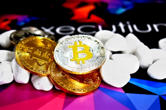 Several physical Bitcoin coins, displaying their iconic 'B' symbol, are placed on a colorful background with abstract geometric shapes. The coins are surrounded by smooth, white stones or pebbles, adding texture and contrast to the image.