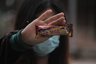 A hand holding a Beng-Beng wafer snack, partially obscuring the person's face, which is also covered with a blue mask. The snack wrapper is red and yellow, featuring images and text indicating it is a chocolate-flavored treat. The background is blurred, emphasizing the hand and the snack bar.