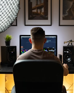 A person is sitting at a desk facing a large computer monitor, likely working on video or photo editing. The setup includes speakers on either side of the monitor and a potted plant. The background displays framed pictures of urban scenes, and a bright, soft light illuminates the space.