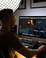 A person is sitting at a computer desk working on a video editing project. The workspace is illuminated by soft lighting from a large grid light panel. The computer screen displays a video editing software interface with various color grading tools and a small video preview. The person appears focused on their task.