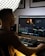 A person is sitting at a computer desk working on a video editing project. The workspace is illuminated by soft lighting from a large grid light panel. The computer screen displays a video editing software interface with various color grading tools and a small video preview. The person appears focused on their task.