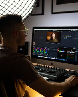 A person is sitting at a computer desk working on a video editing project. The workspace is illuminated by soft lighting from a large grid light panel. The computer screen displays a video editing software interface with various color grading tools and a small video preview. The person appears focused on their task.