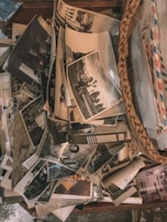 A collection of old black and white photographs is scattered across a wooden surface next to a woven basket containing envelopes. The images appear to depict various scenes, possibly family moments, with some showing people in outdoor settings.