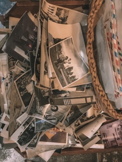 A collection of old black and white photographs is scattered across a wooden surface next to a woven basket containing envelopes. The images appear to depict various scenes, possibly family moments, with some showing people in outdoor settings.
