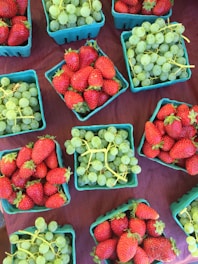 Brightly colored cartons filled with fresh strawberries and green grapes are arranged on a dark surface, creating a vibrant display. Each green carton is neatly packed with ripe, juicy strawberries or bunches of green grapes, showcasing a bountiful collection of fruit.